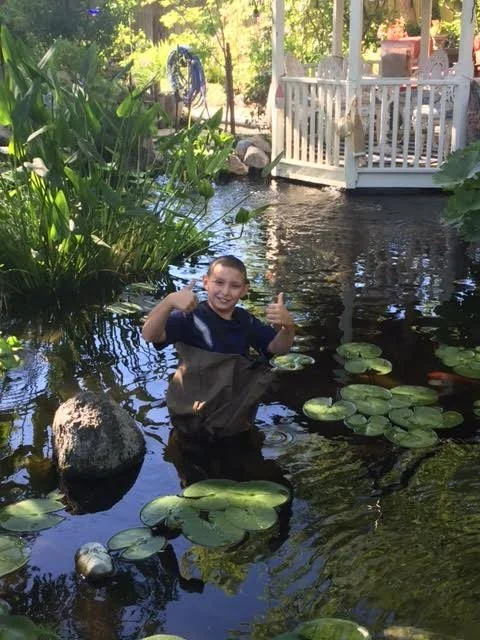 A young crew member in waders giving two thumbs-up from the middle of a lily pond