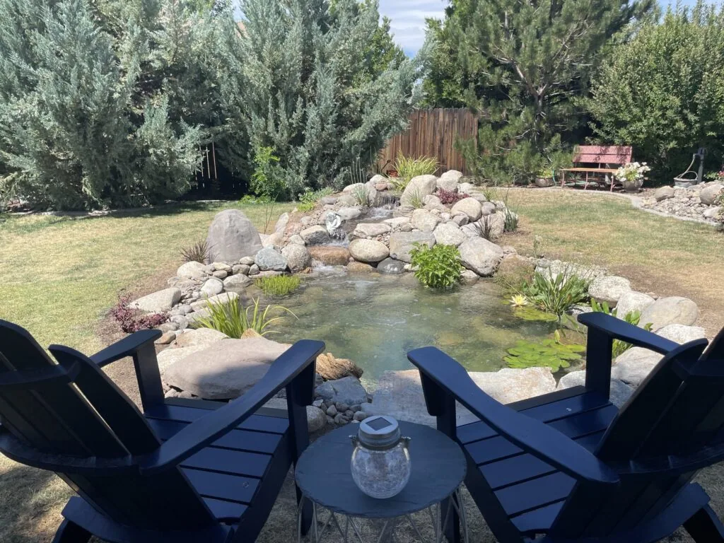 Two navy Adirondack chairs facing a finished backyard pond with a boulder edge and water plants