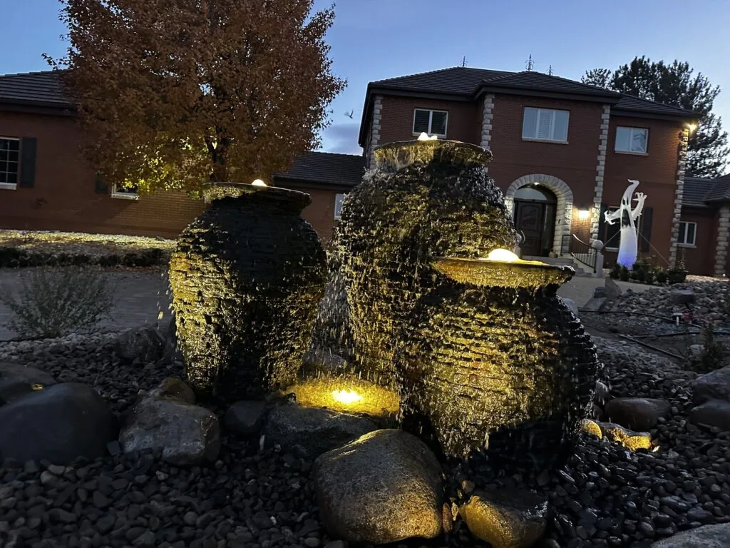 Three stacked-slate urn fountains glowing with landscape lights at dusk in front of a brick house